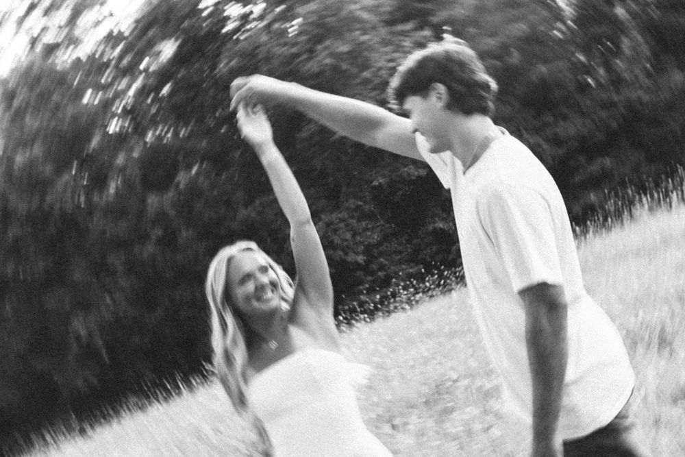 A black and white photo of a couple holding hands and spinning together outdoors in a playful moment.
