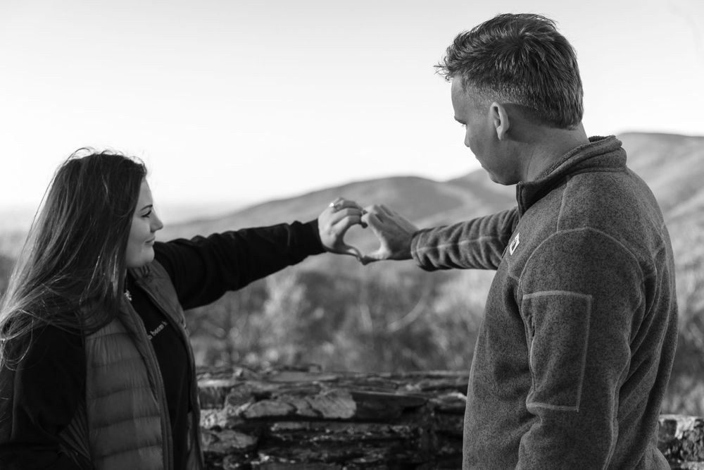 During a proposal the couple creates a heart with their hands while standing on mountain overlook.
