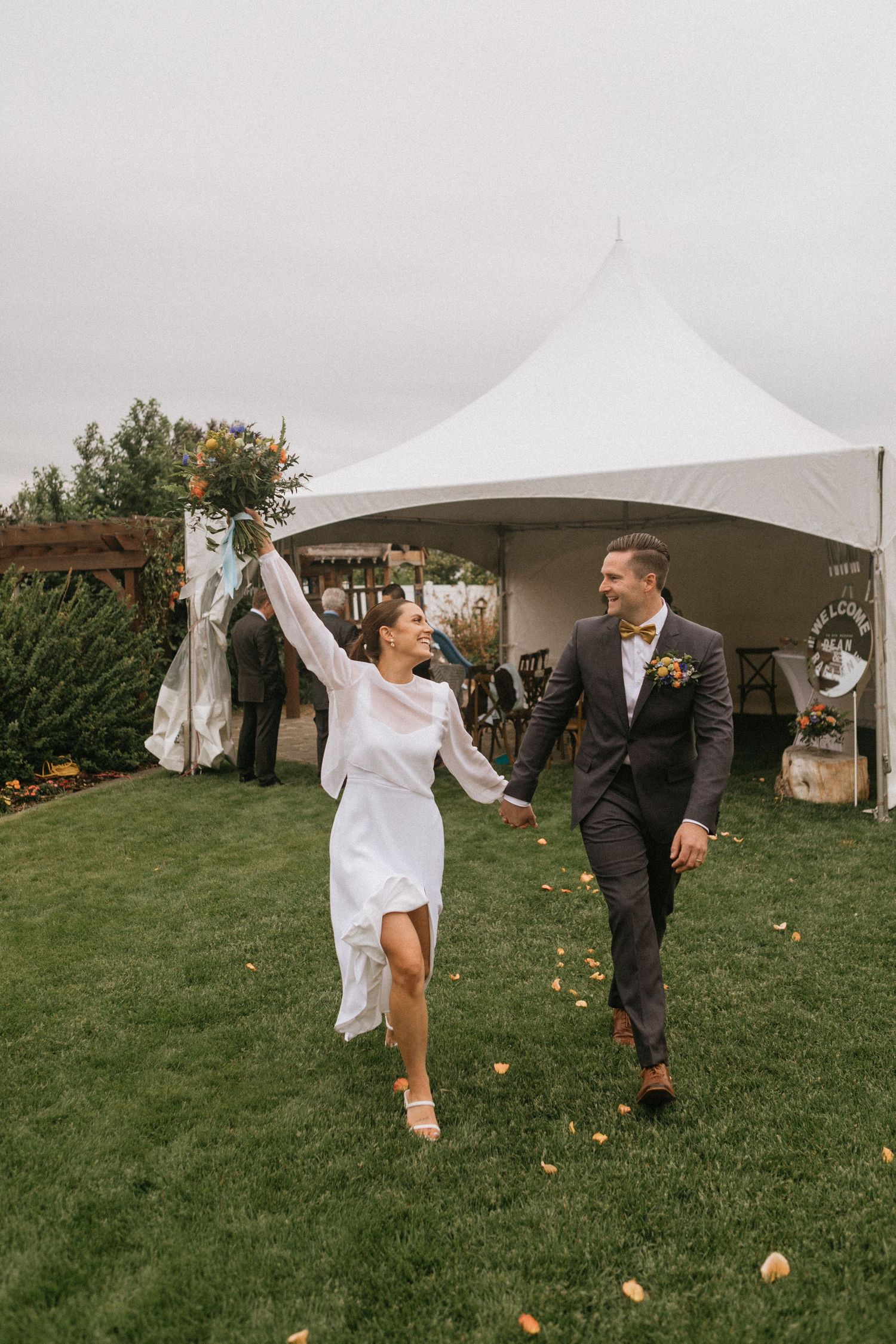Joyful couple running on grass beneath white tent during outdoor wedding celebration.
