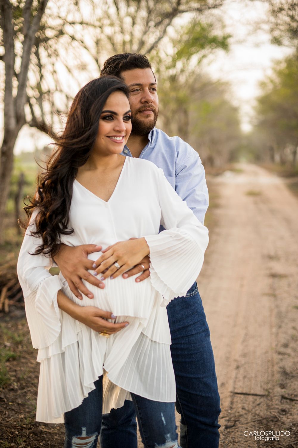 Couple in white and blue attire embrace on a rustic country road during maternity shoot.