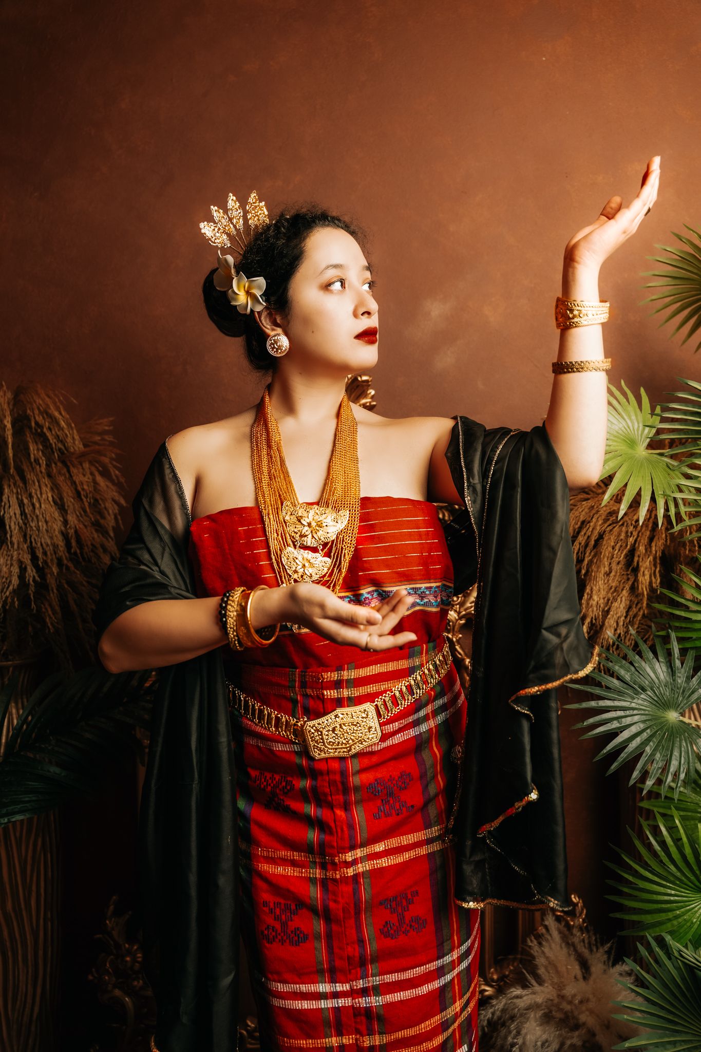 Traditional Southeast Asian fashion portrait featuring ornate red dress and gold jewelry against dark background.