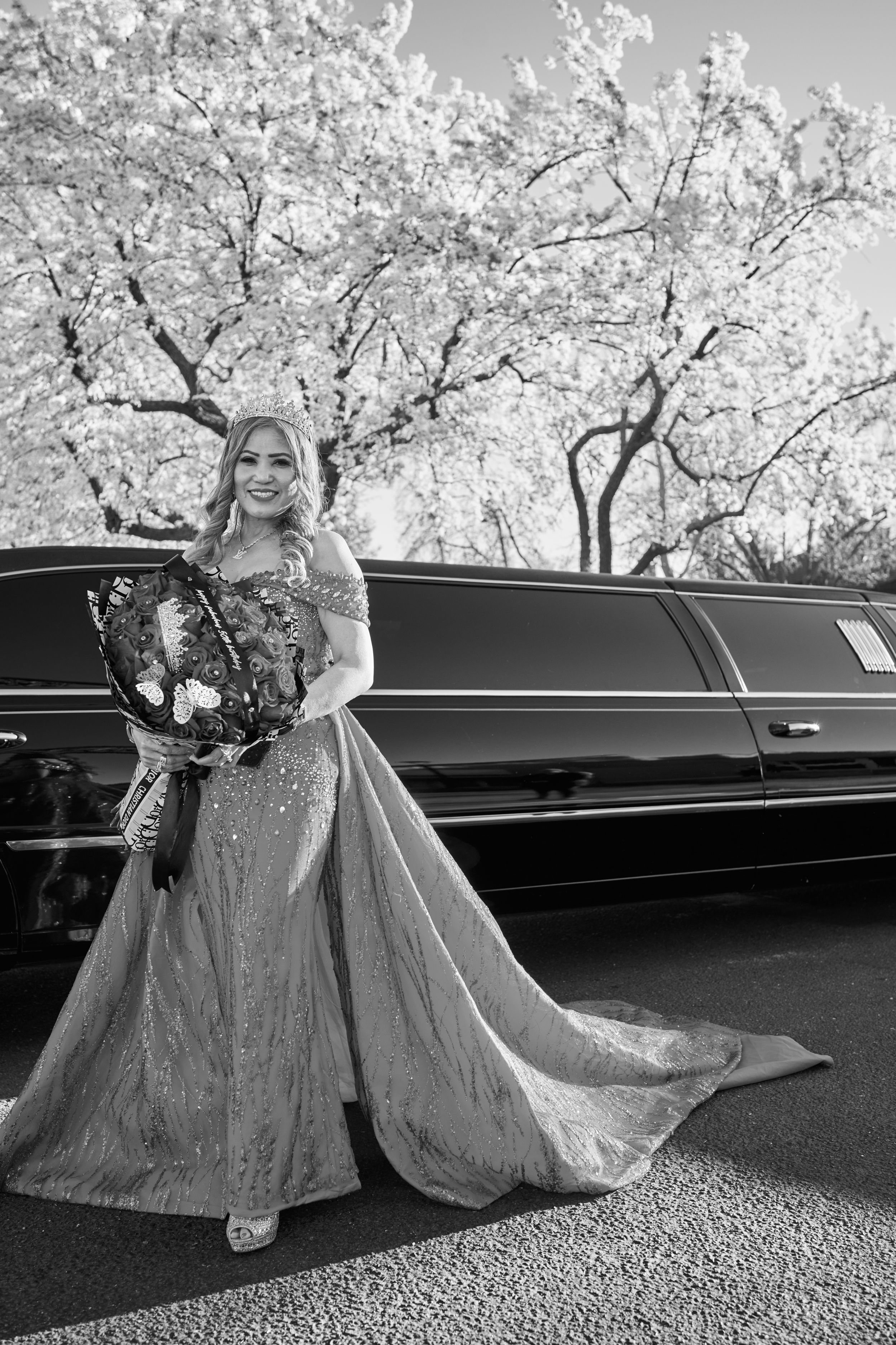 Black and white photo of a bride in a sparkly gown standing beside a luxury stretch limousine under flowering spring trees.