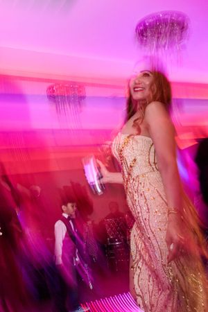 A glamorous celebration scene at an event with pink and purple lighting showing someone dancing in a sparkly gold dress.
