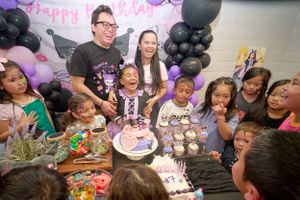 Children celebrate at a birthday party with black and pink balloon decorations as they gather around a cake.
