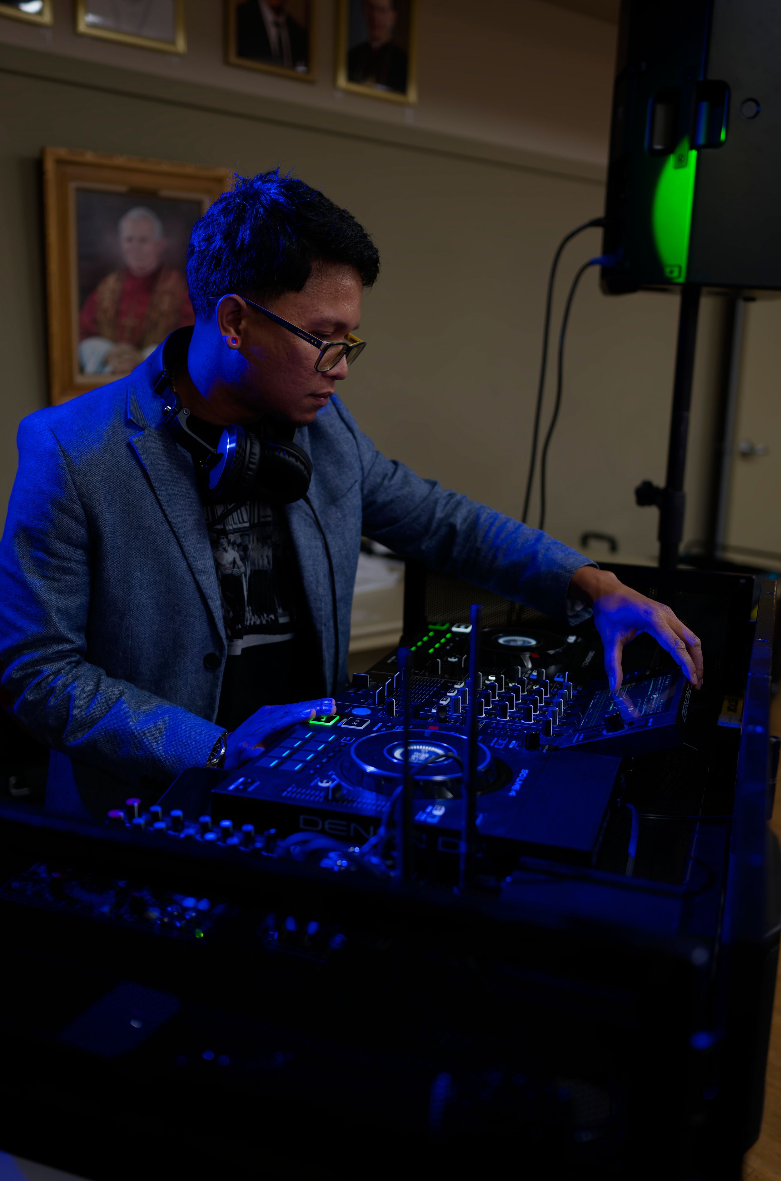 A DJ performs at night under blue lighting while operating professional mixing equipment and wearing a blazer.
