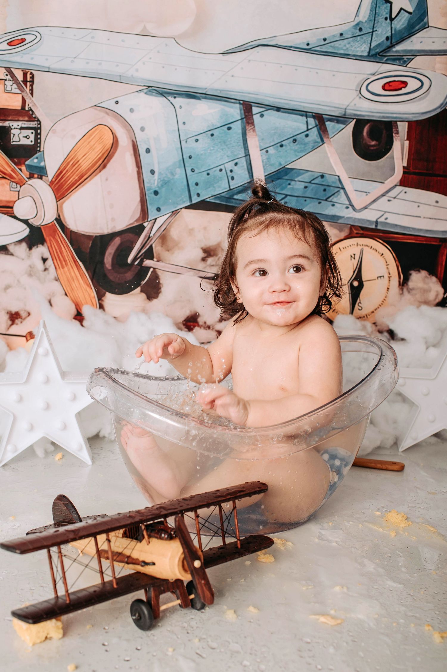 A baby sits in a clear container surrounded by vintage aviation props and clouds in a styled photo shoot.