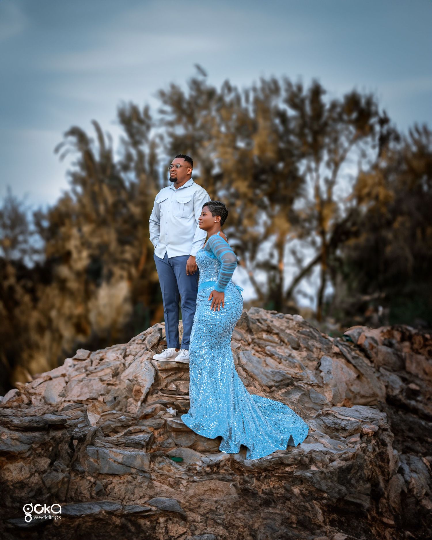 Two people stand on a rocky outcrop, one wearing jeans and white top, the other in a flowing blue formal dress.