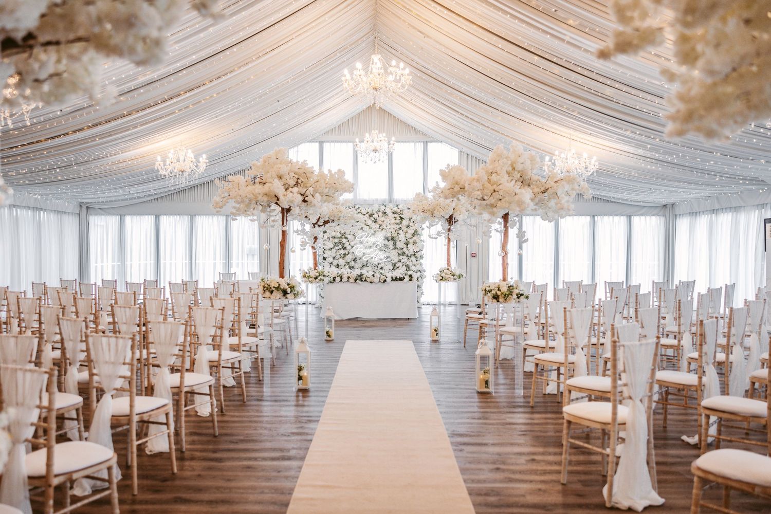 Elegant all-white wedding reception setup with crystal chandeliers and draped fabric ceiling in a spacious ballroom.