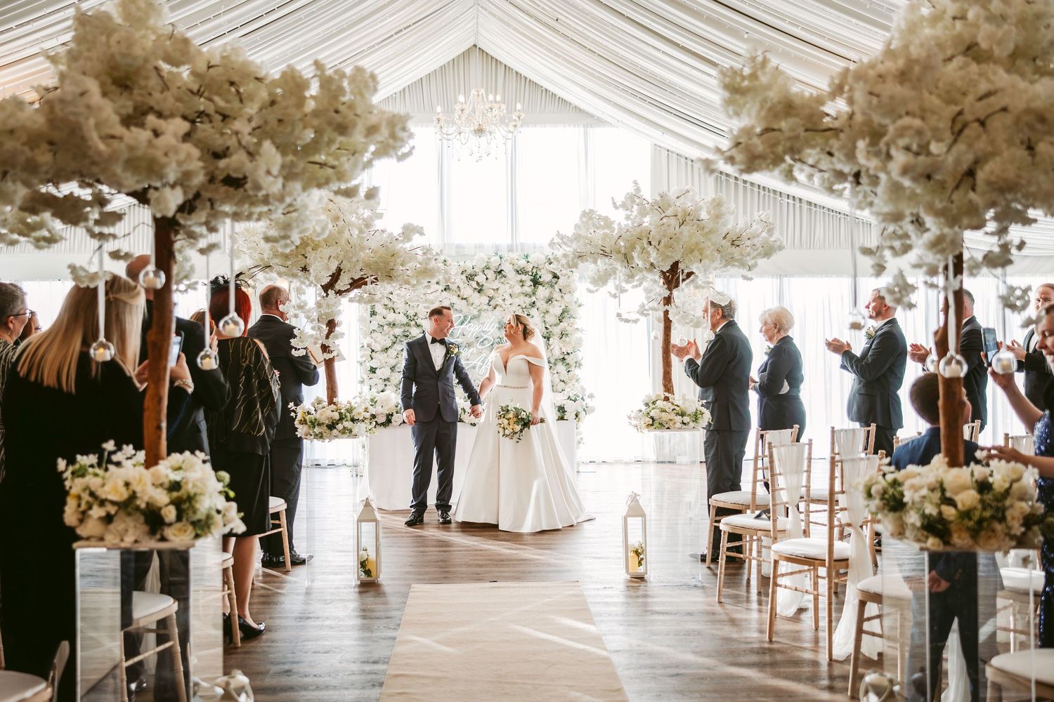 A romantic wedding ceremony takes place in a bright greenhouse venue decorated with trees and floral arrangements.