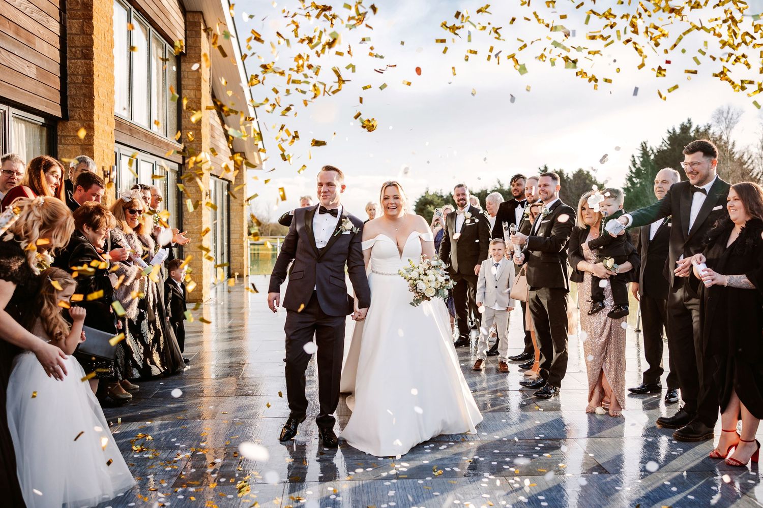 Wedding guests toss golden autumn leaves during a joyful celebration outside a venue with large windows.