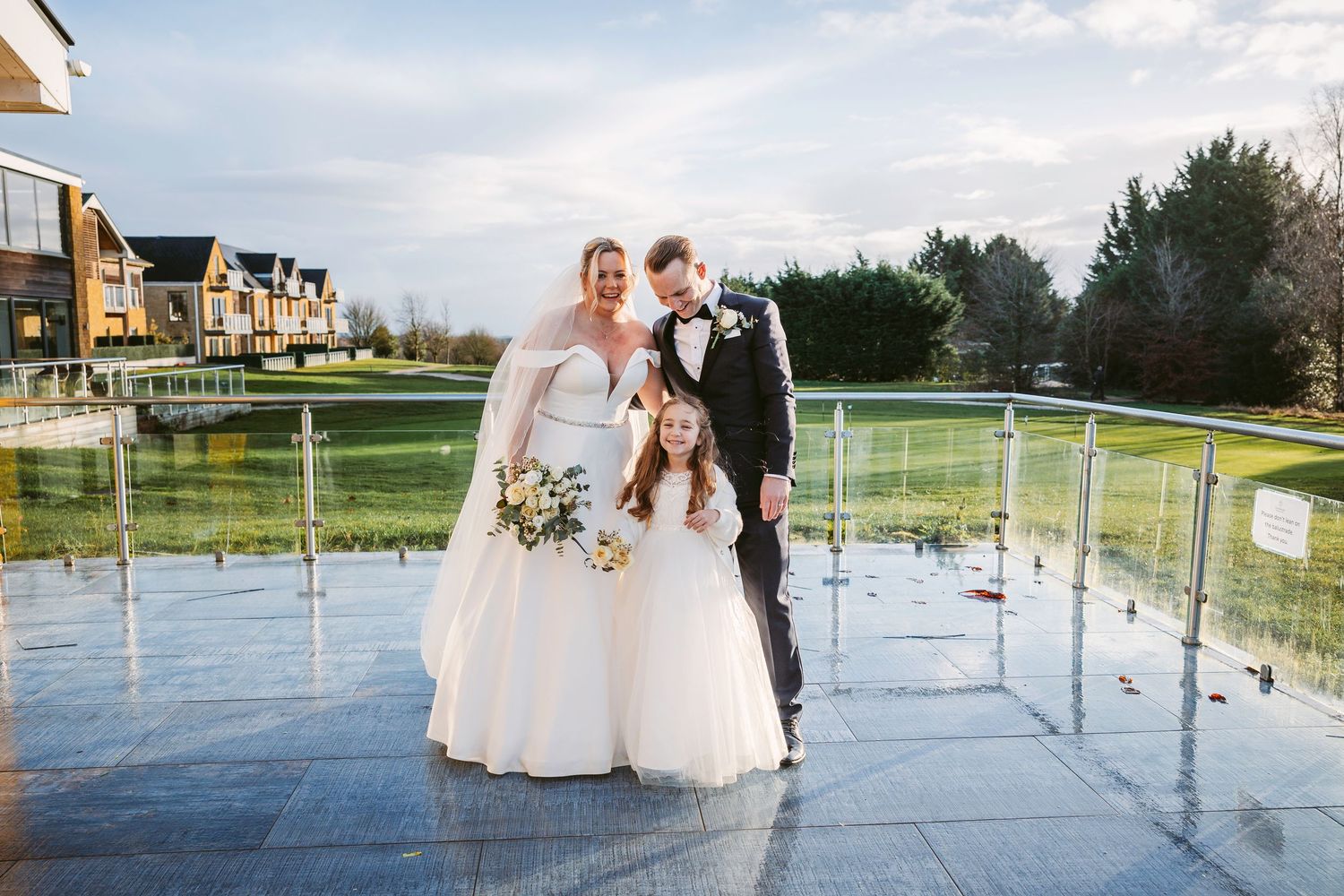 A wedding couple poses with their dog on a glass balcony overlooking a golf course on a cloudy day.