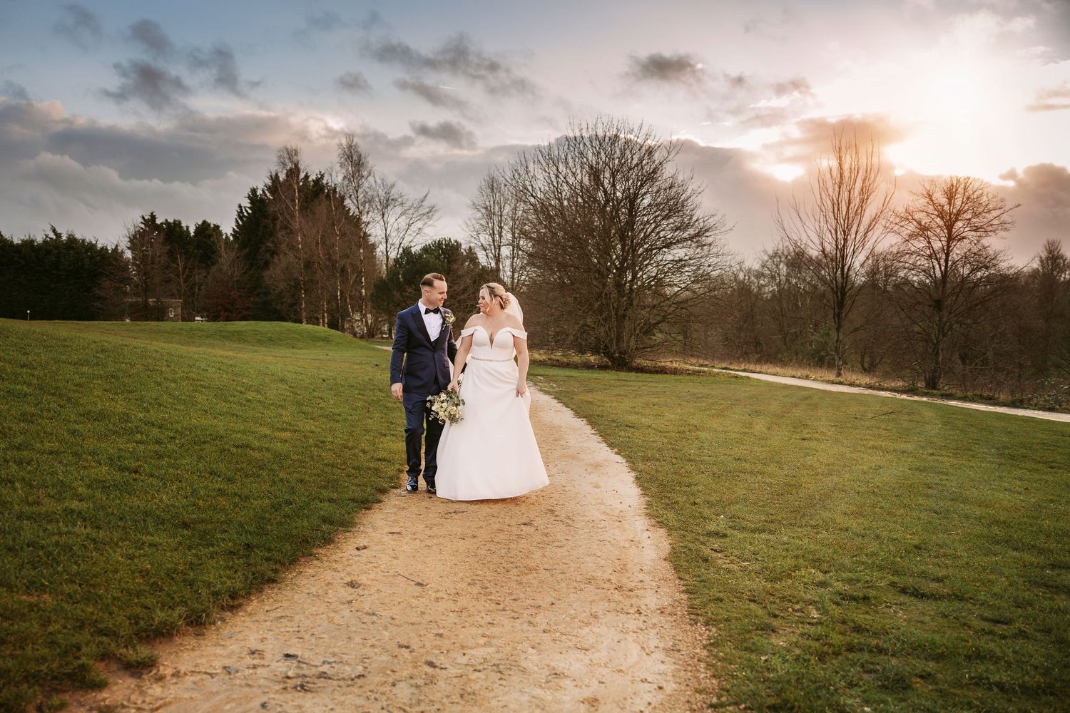 A couple walks along a winding path through a scenic countryside at sunset with dramatic clouds in the sky.