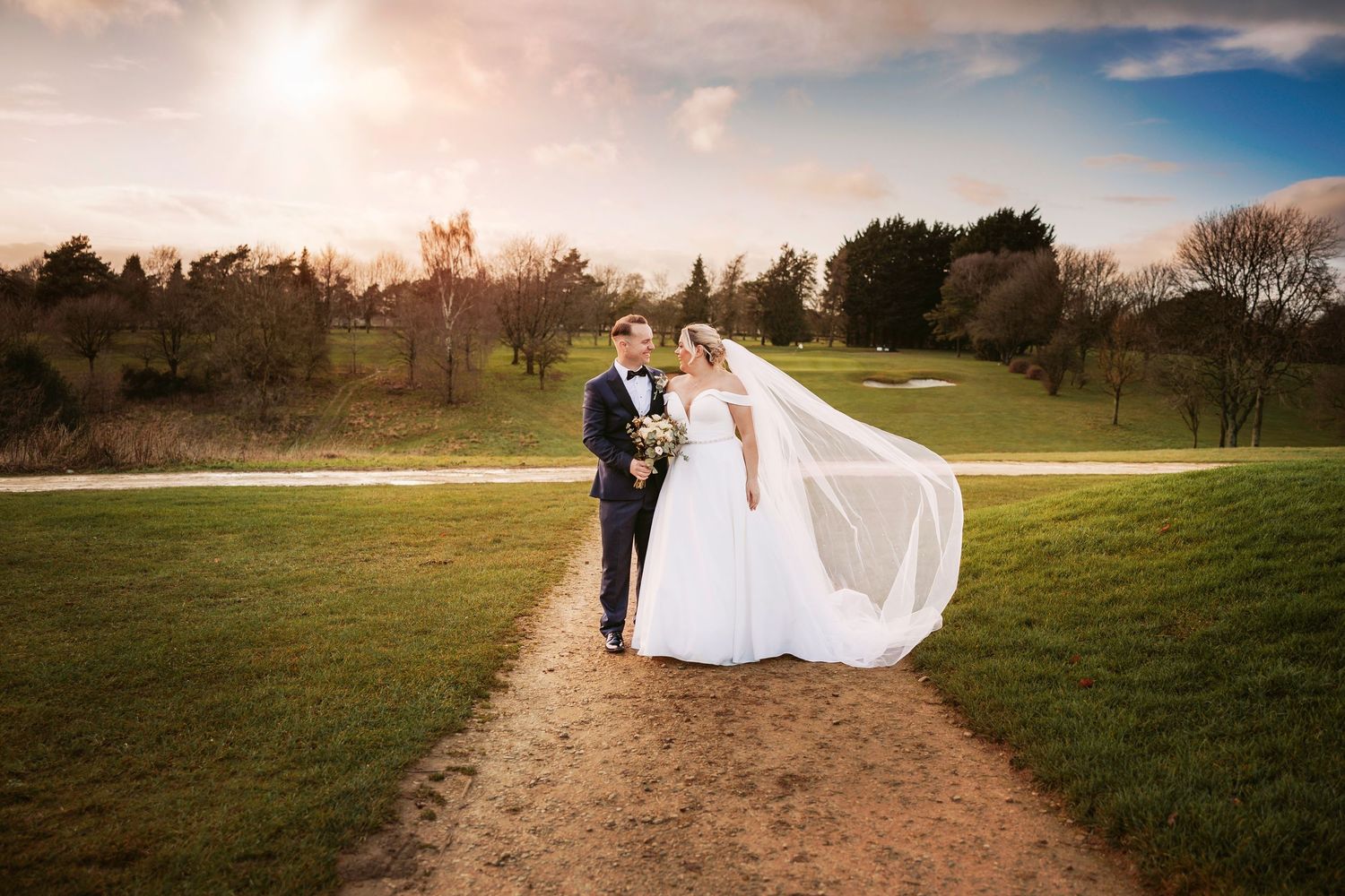 A couple in wedding attire walks down a winding dirt path through a grassy field at sunset.