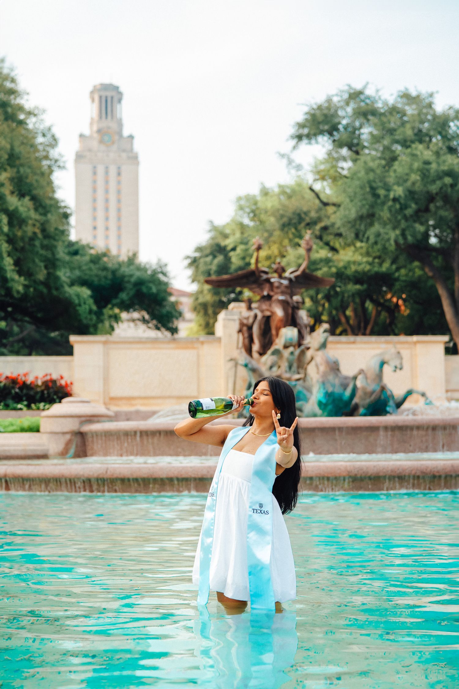 A person in a white dress splashes in a turquoise fountain pool with a tall university tower visible in the background.
