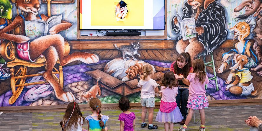 A woman speaking to toddlers in front of the Columbine Library children's mural