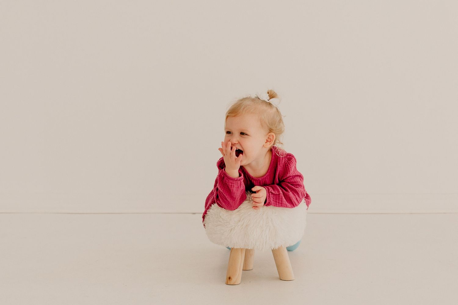 An adorable toddler girl in a pink top sits on a small stool with her hand near her mouth.