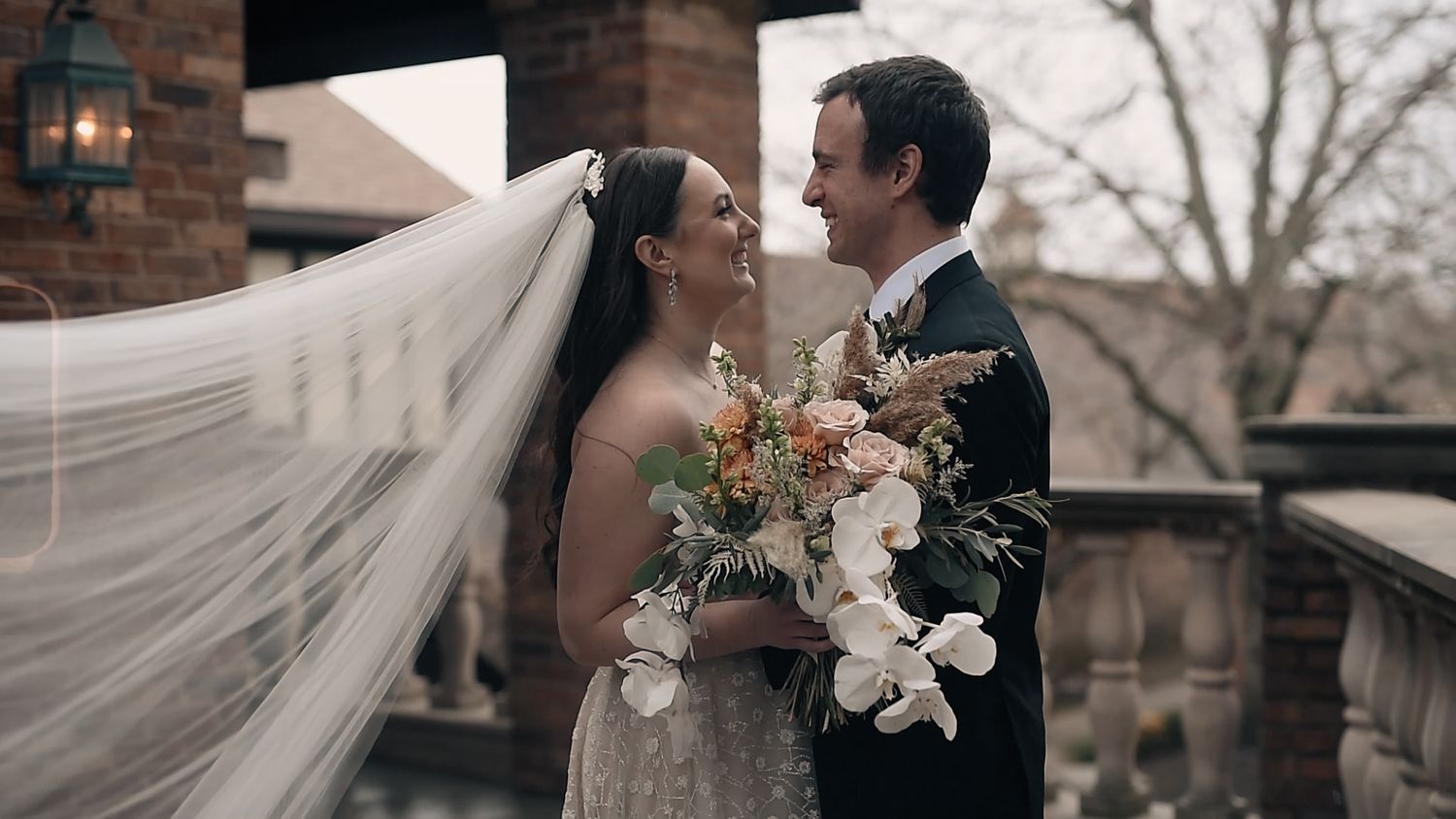 A romantic wedding couple shares a tender moment on a balcony with flowing veil and autumn floral bouquet.