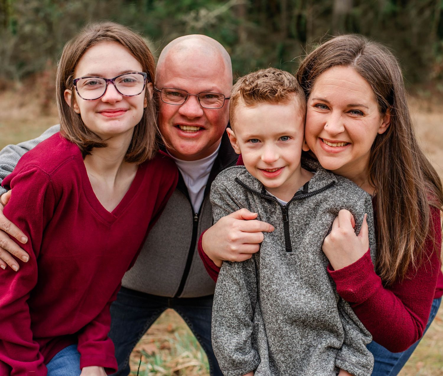 A family in coordinating brown and beige outfits poses together outdoors in a wooded setting.