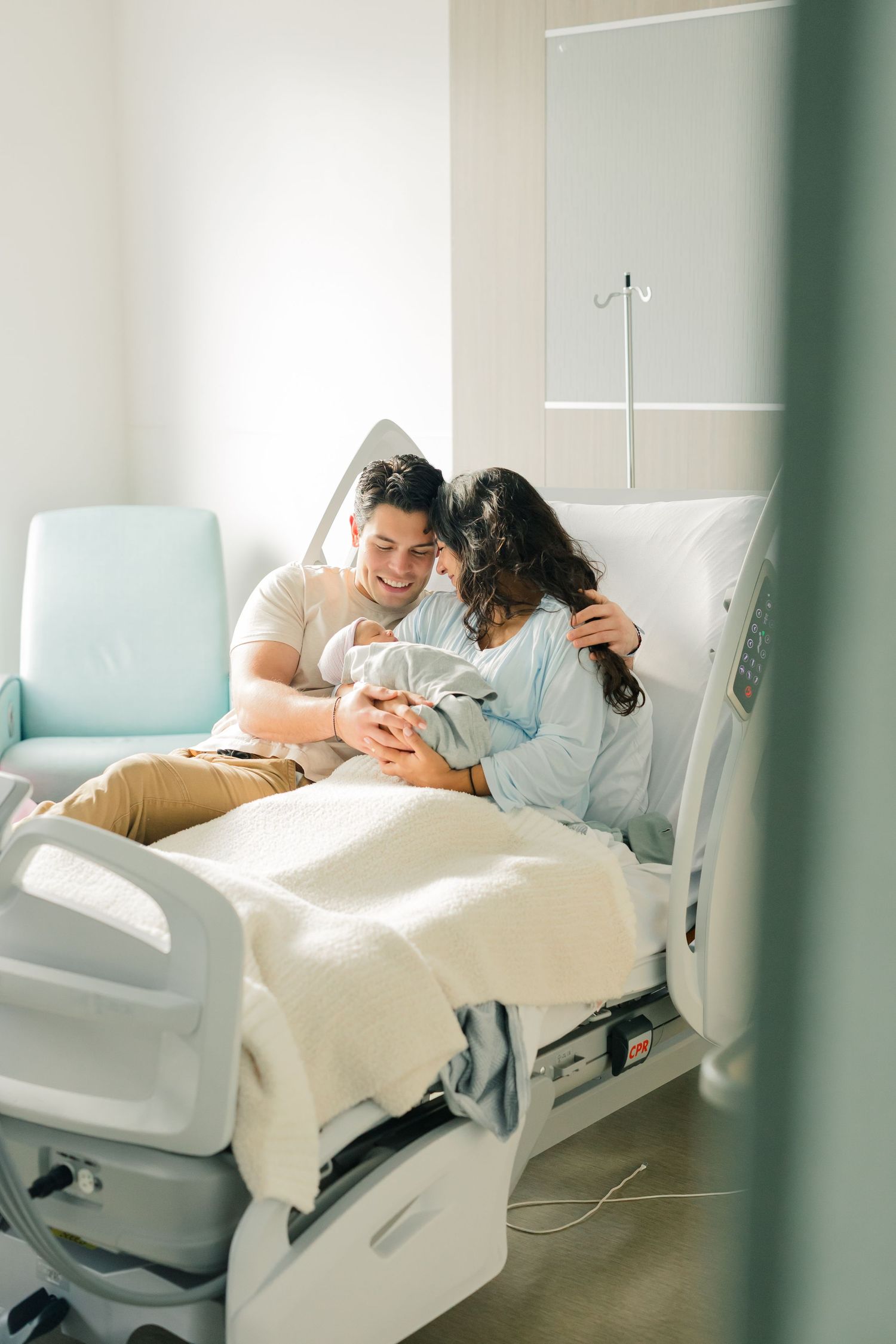 A couple rests together in a hospital room with white bedding and mint green walls.