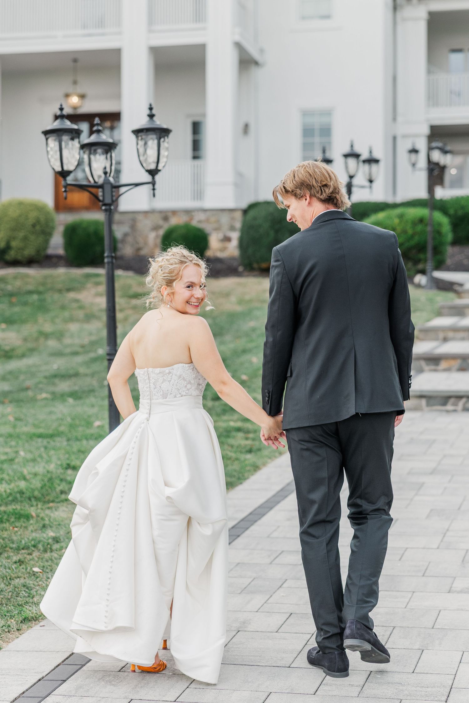 A couple strolls down an elegant stone walkway with lamp posts and landscaping.