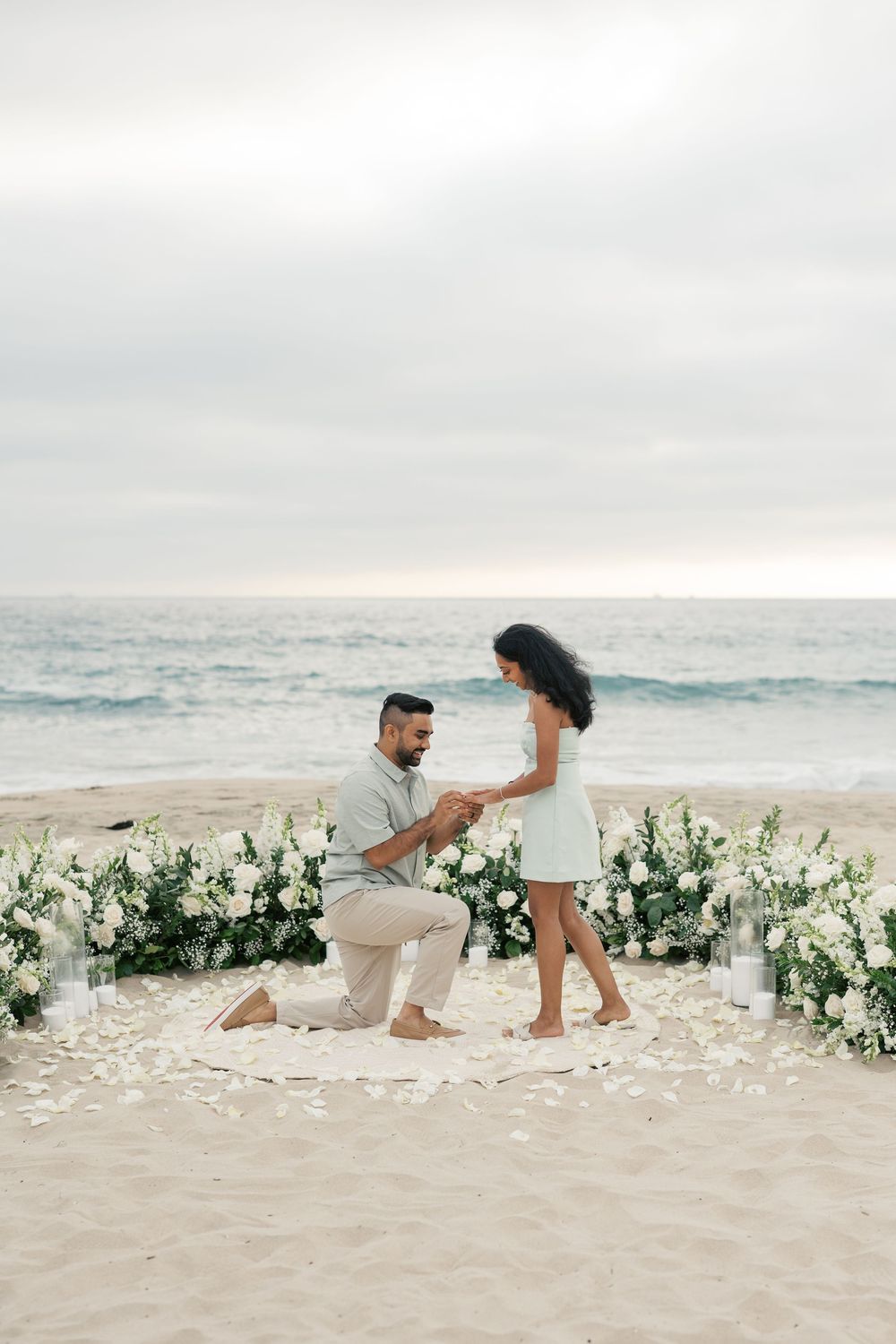 A romantic beach proposal scene with white floral arrangements and rose petals scattered on the sand at sunset.