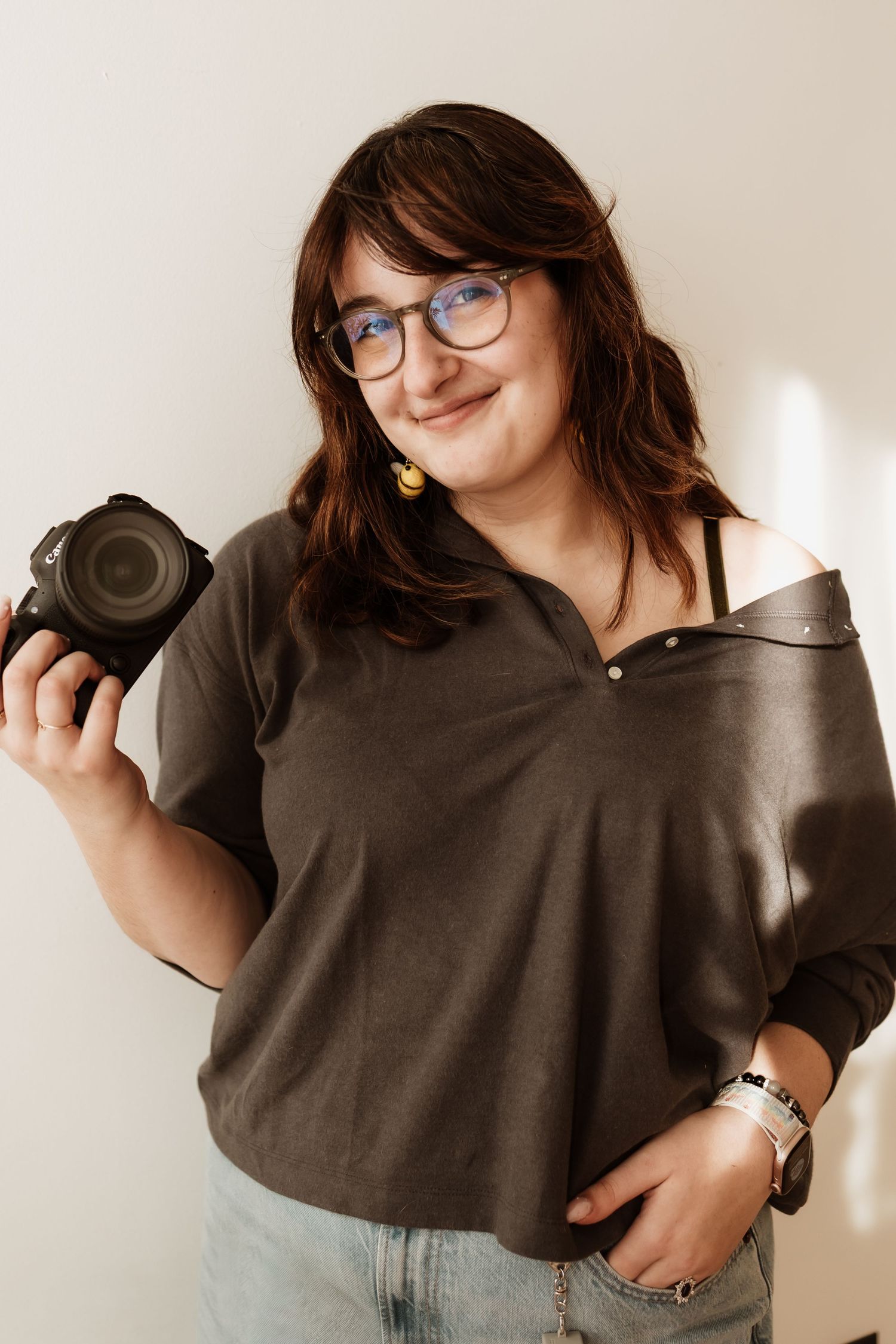 A photographer in a brown sweater poses playfully with camera equipment against a light background.
