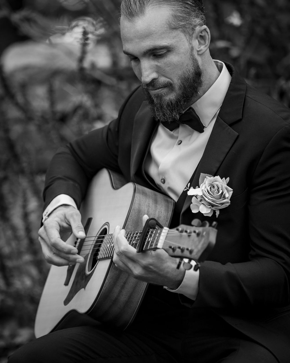 Close-up black and white photo of someone playing an acoustic guitar with a white boutonniere on their formal suit.