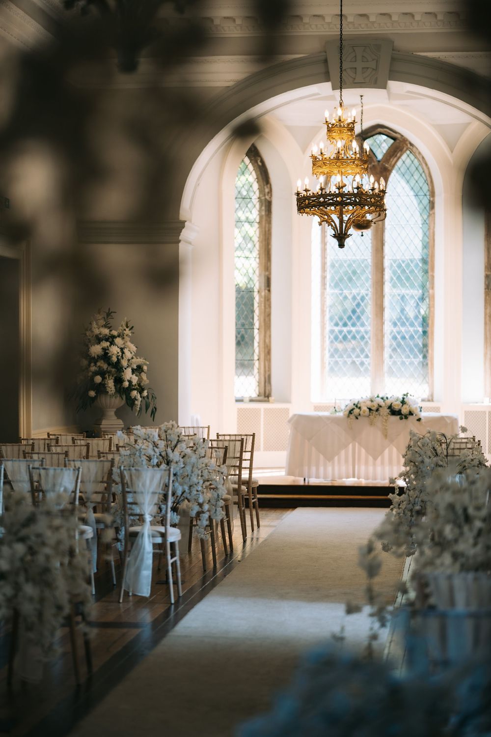 Elegant wedding ceremony setup in a chapel with arched windows, chandelier, and white floral arrangements lining the aisle.