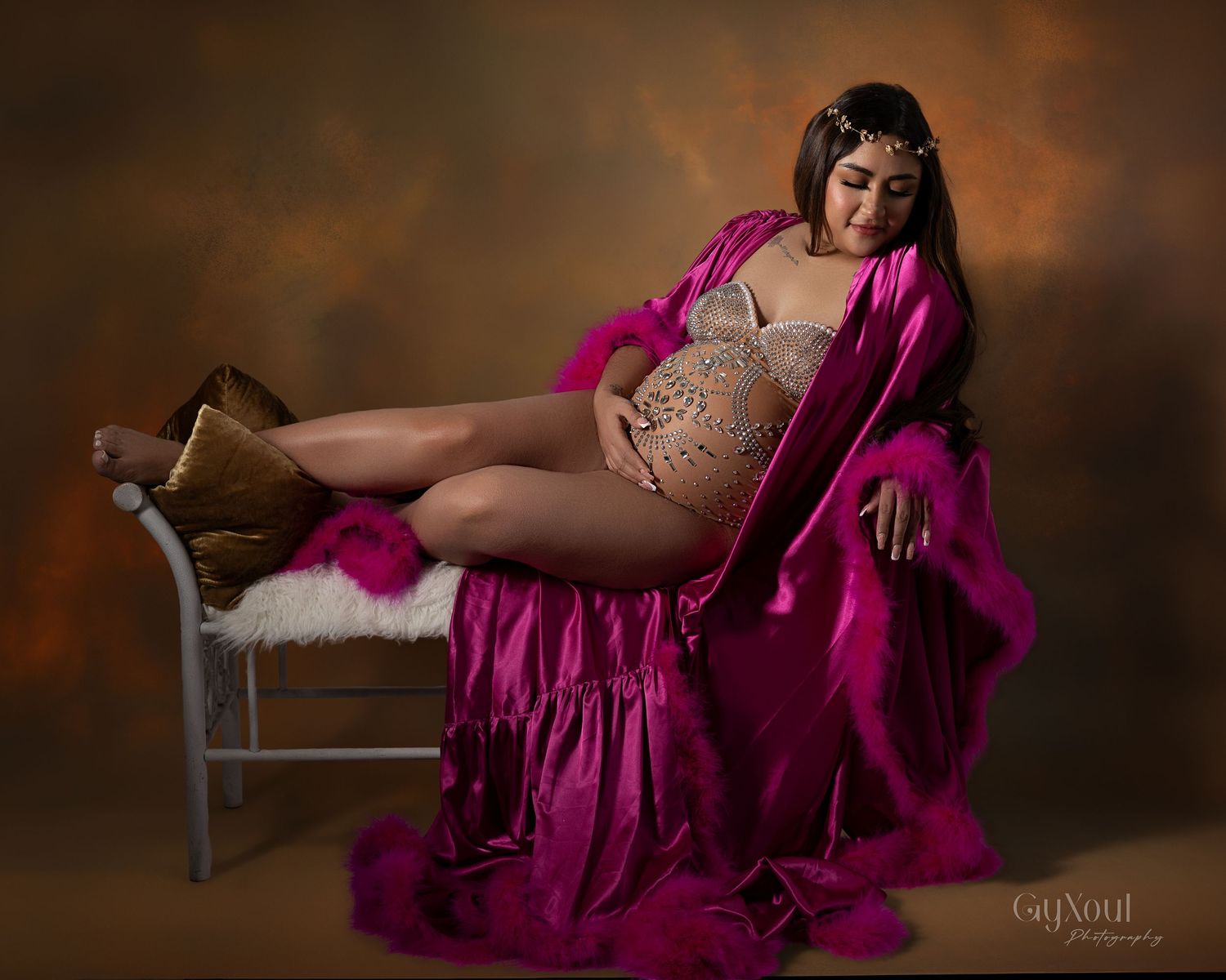 A pregnant woman in a flowing magenta maternity gown poses elegantly against a dark brown backdrop in a studio setting.