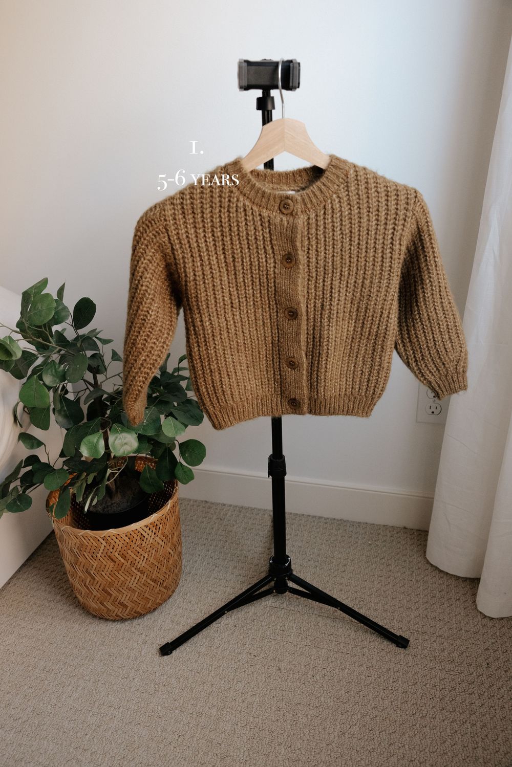Chunky knit tan cardigan sweater displayed on black photography stand with plant decor.