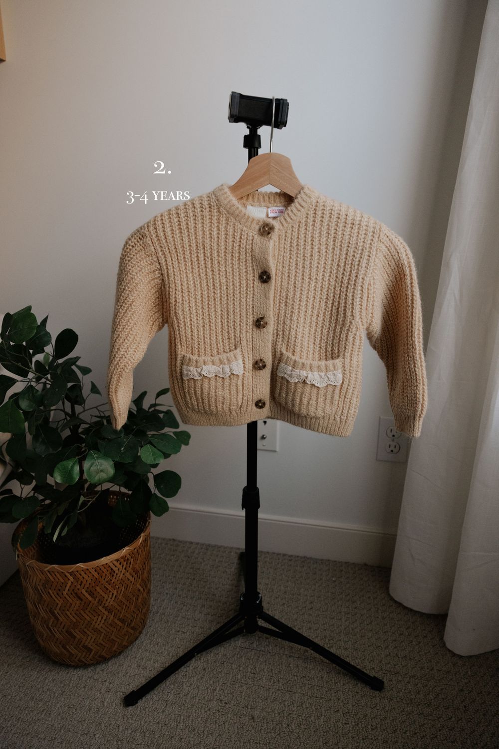 Cream colored baby cardigan with decorative pockets displayed on stand beside potted plant.