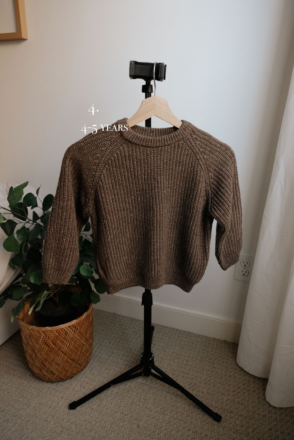 Brown knit sweater displayed on black photography stand with woven basket and plant.