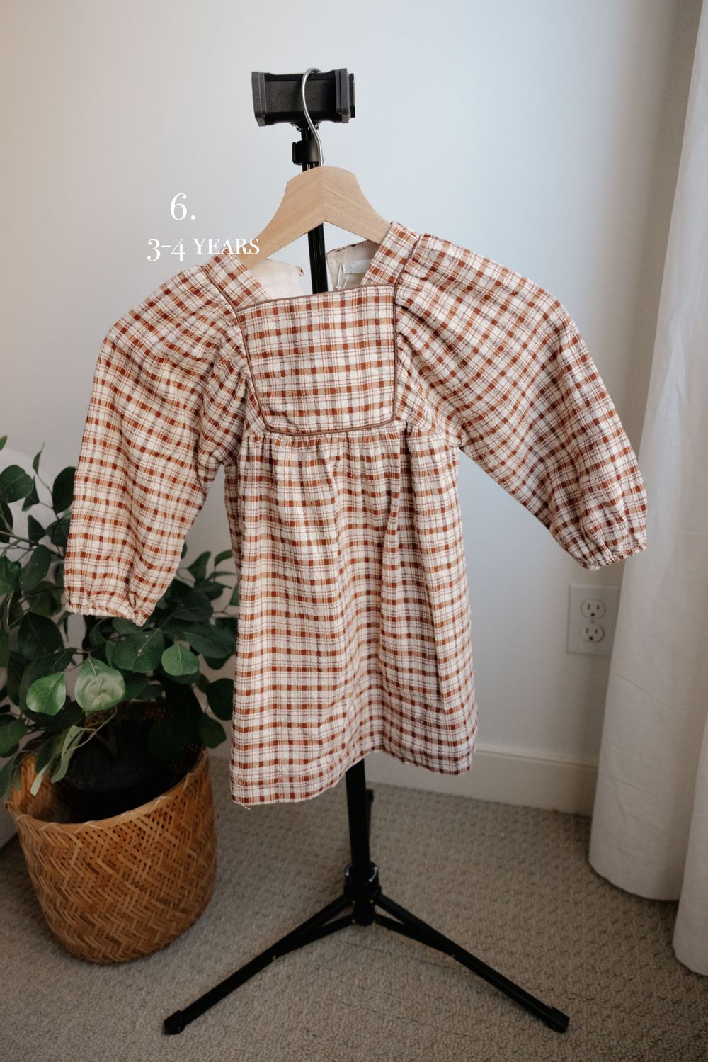 Vintage-style checkered dress with puff sleeves displayed on stand next to potted plant.