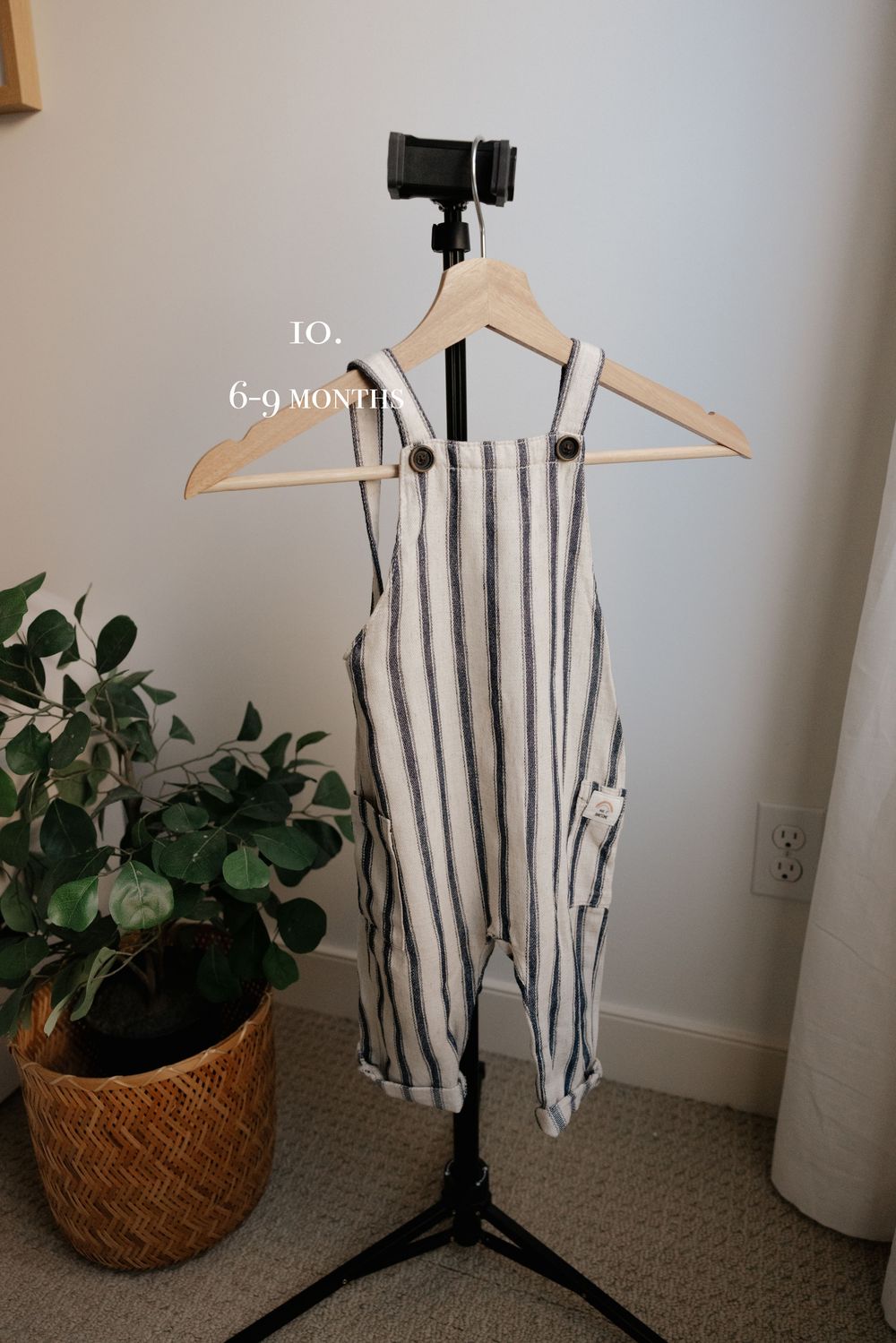 White and black striped baby romper hanging on wooden hanger against light wall with potted plant.