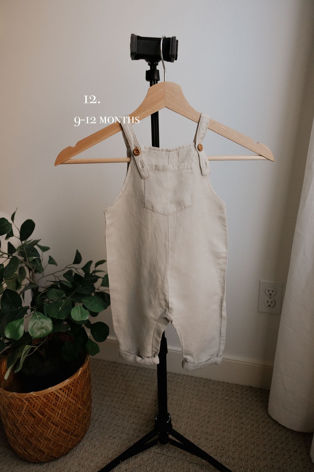 Beige linen baby shortalls displayed on wooden hanger against neutral wall.