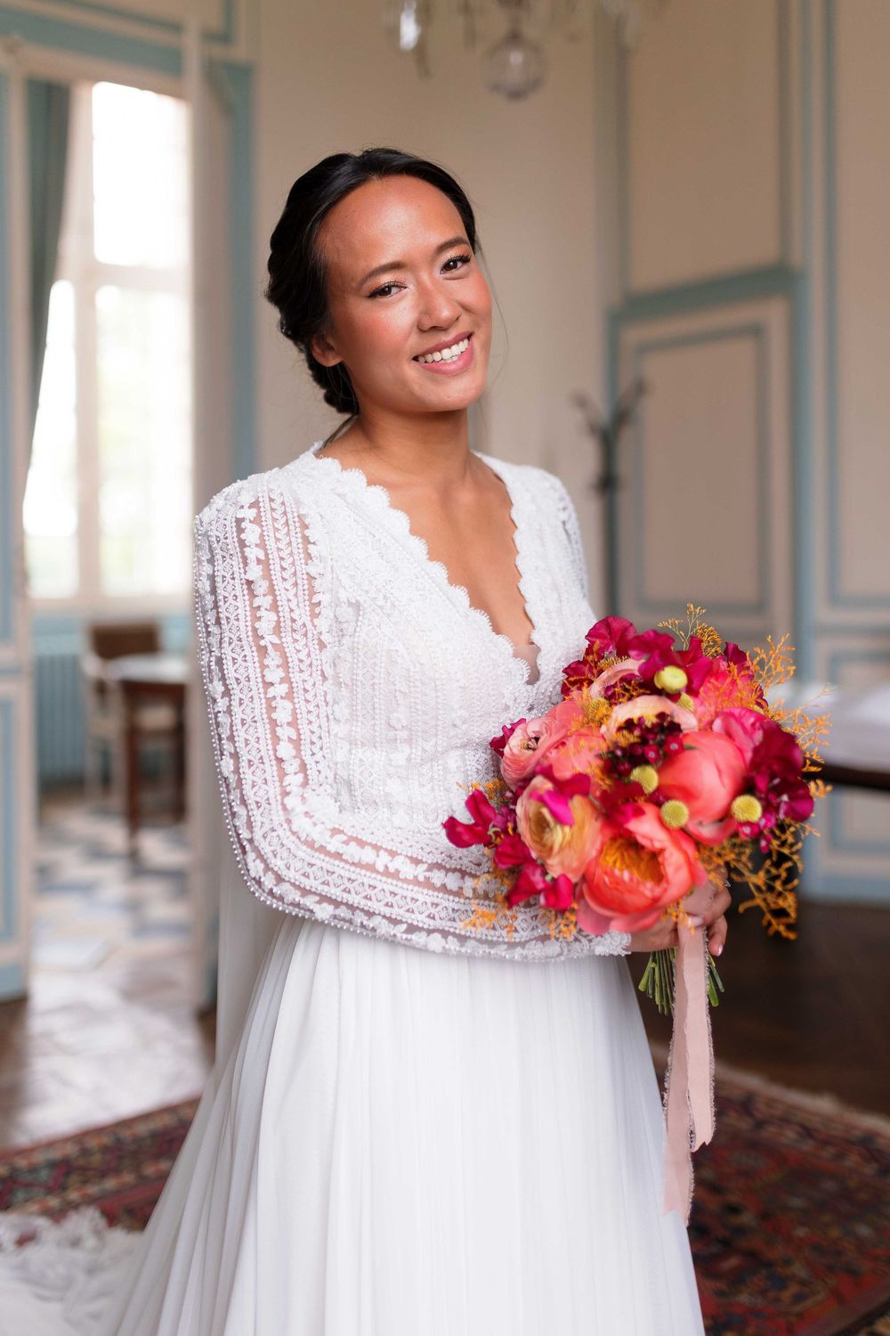 A bride in a white empire waist dress holds a vibrant coral and pink bouquet.
