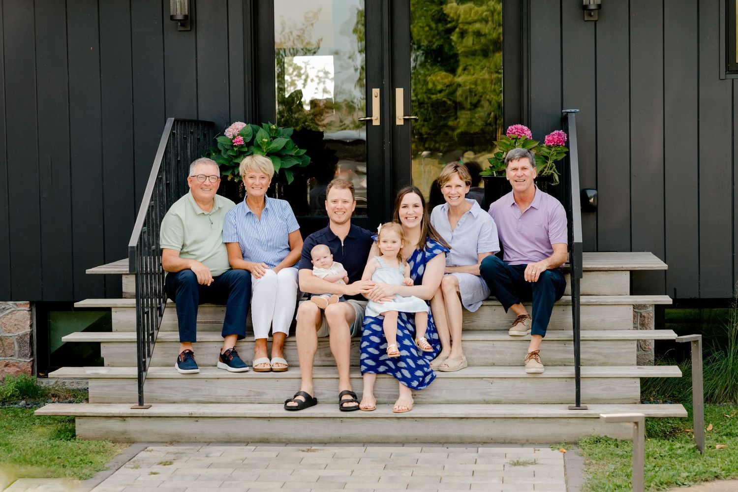 Multi-generational family smiling together on front porch steps of a dark home on a sunny day.
