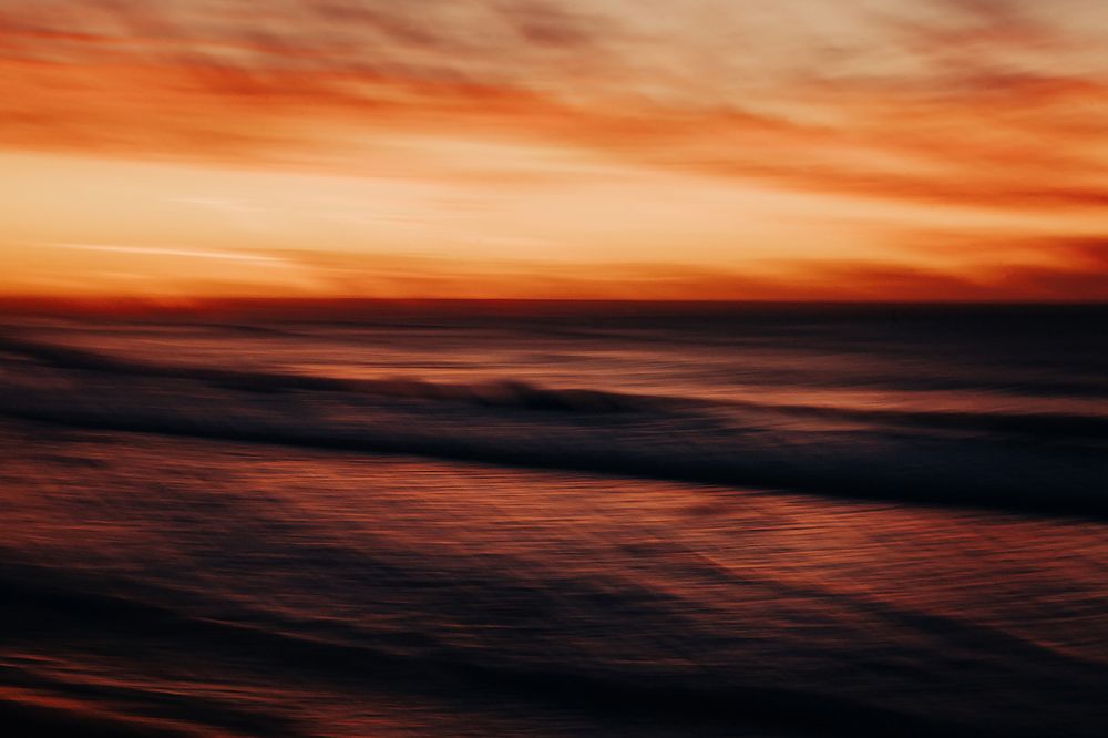 Dark ocean waves roll beneath a vibrant orange and red sunset sky in a moody seascape.