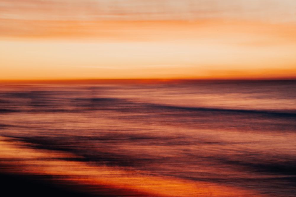A dramatic panoramic seascape at sunset with vibrant orange and purple hues reflecting off the ocean waves.