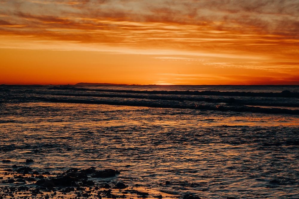Dramatic orange sunset over dark ocean waves with vibrant cloudy sky reflecting on the rippling water surface.
