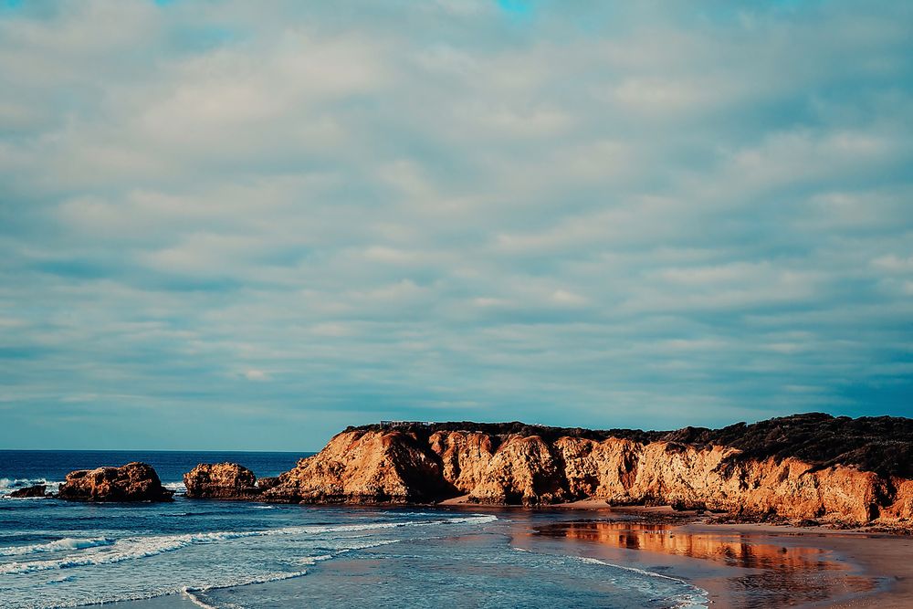 Golden sunlit coastal cliffs rise above a sandy beach with crashing waves under a cloudy turquoise sky.