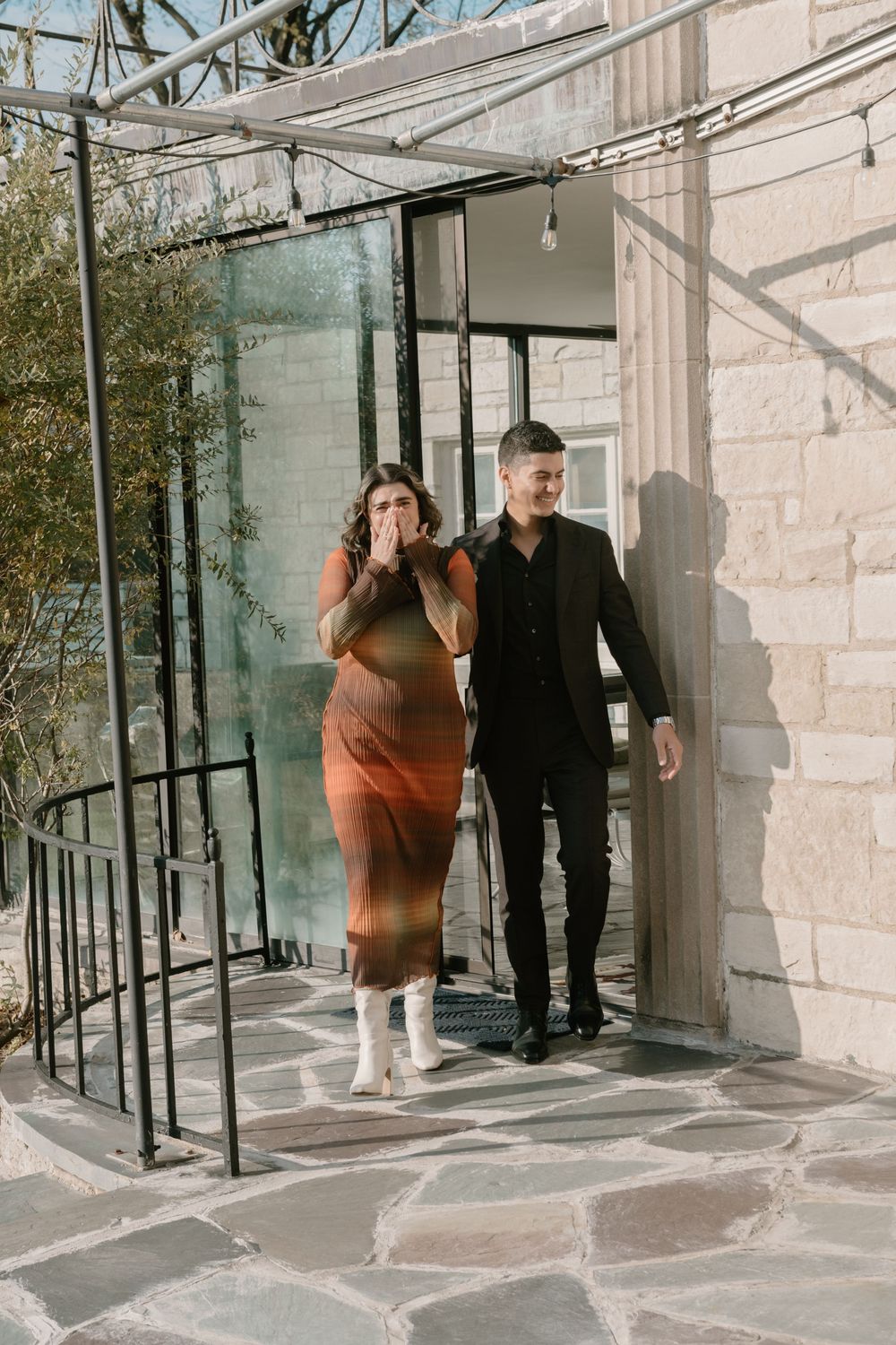 Two people walk along a stone-paved patio with glass walls and natural lighting creating an elegant architectural scene.