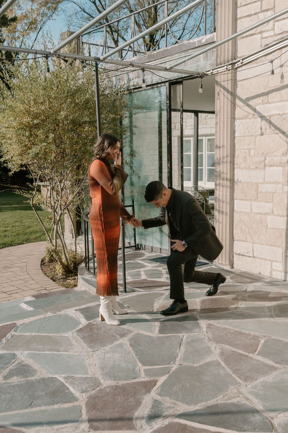 A couple performs a playful dance sequence on a stone patio outside a modern building with glass windows.