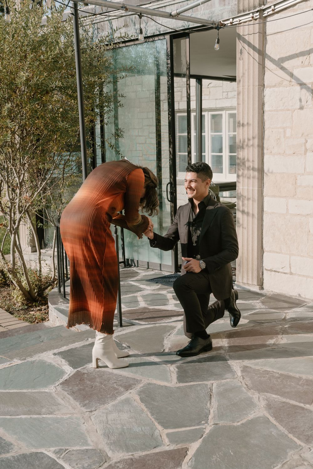 A couple sharing a special moment on a stone patio with glass windows and greenery in the background.