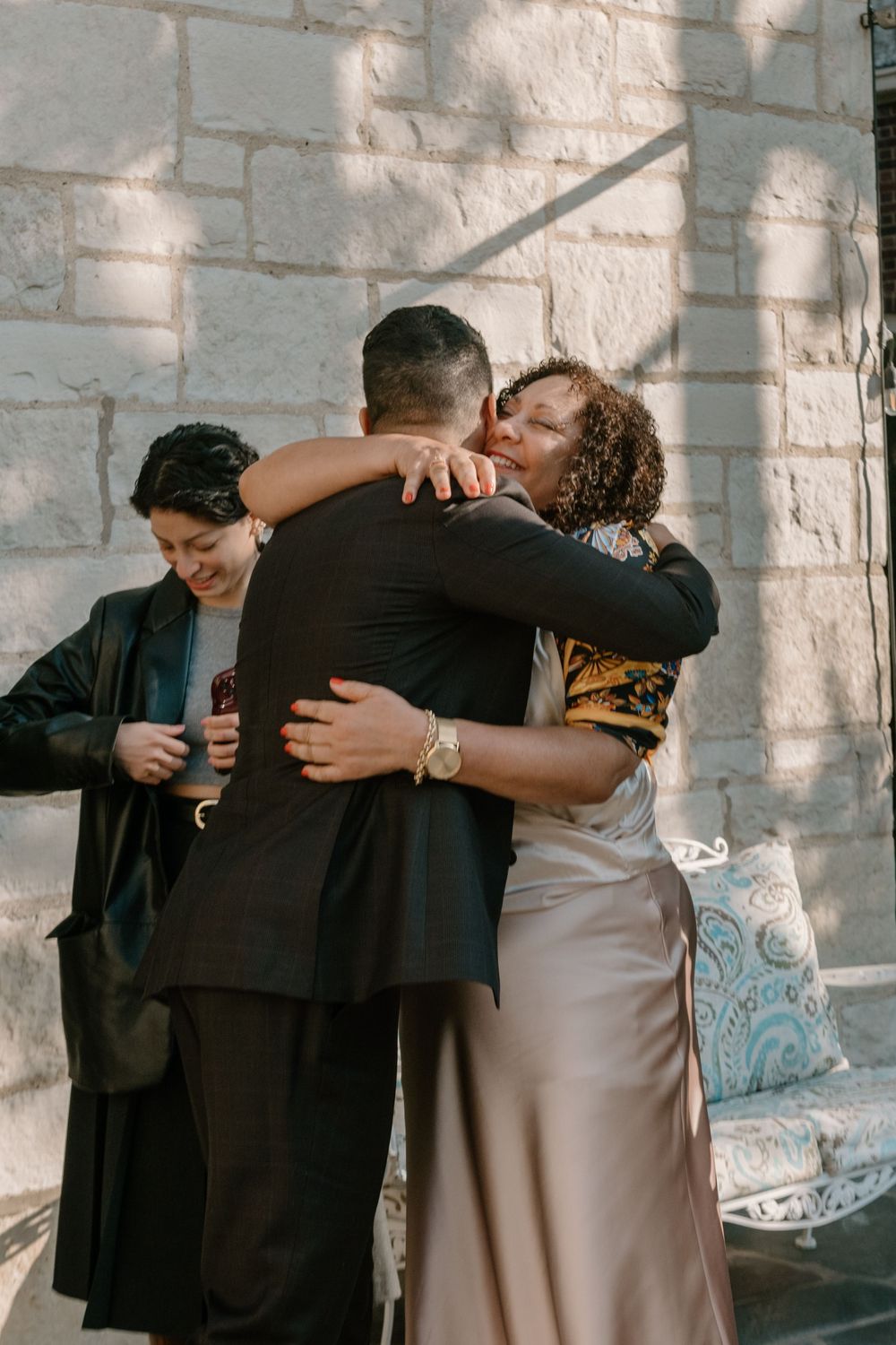 A warm embrace shared between three people against a white stone wall with dappled sunlight.
