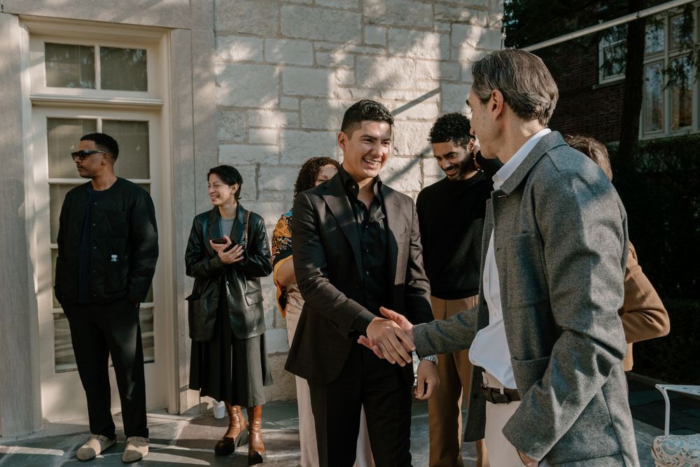 A group of people in formal attire gather and socialize at an outdoor event near a brick building.