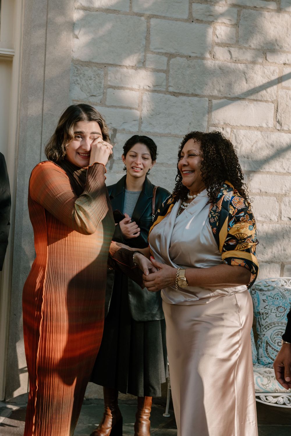 Three people sharing joyful laughter and conversation while standing against a white stone wall outdoors.