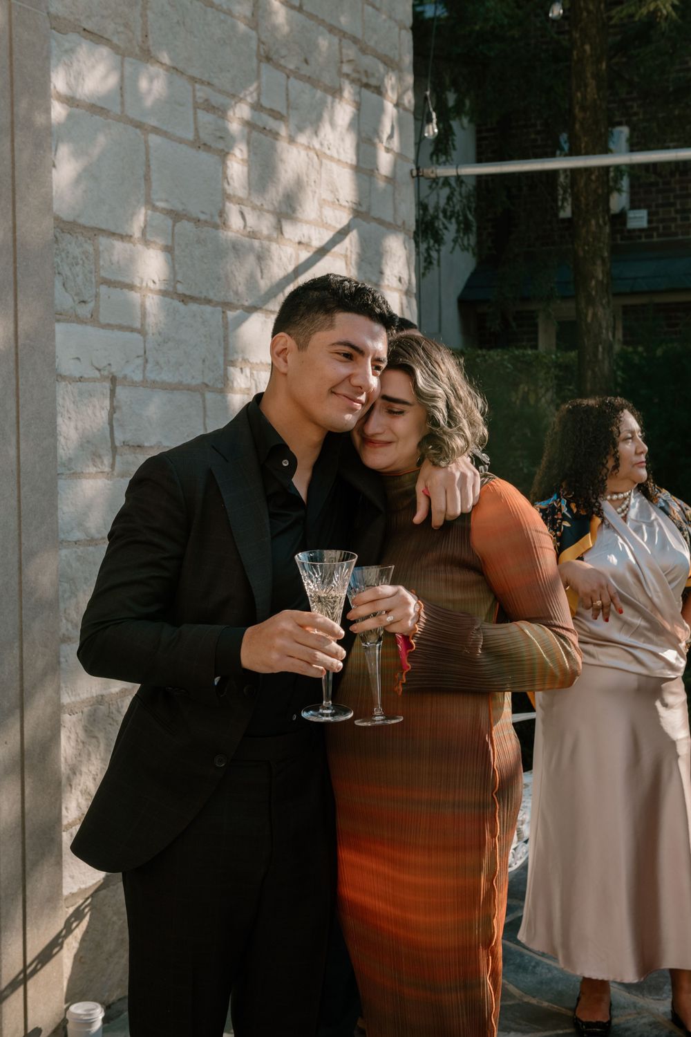 A couple shares an intimate moment while holding drinks at an outdoor gathering with stone walls in the background.