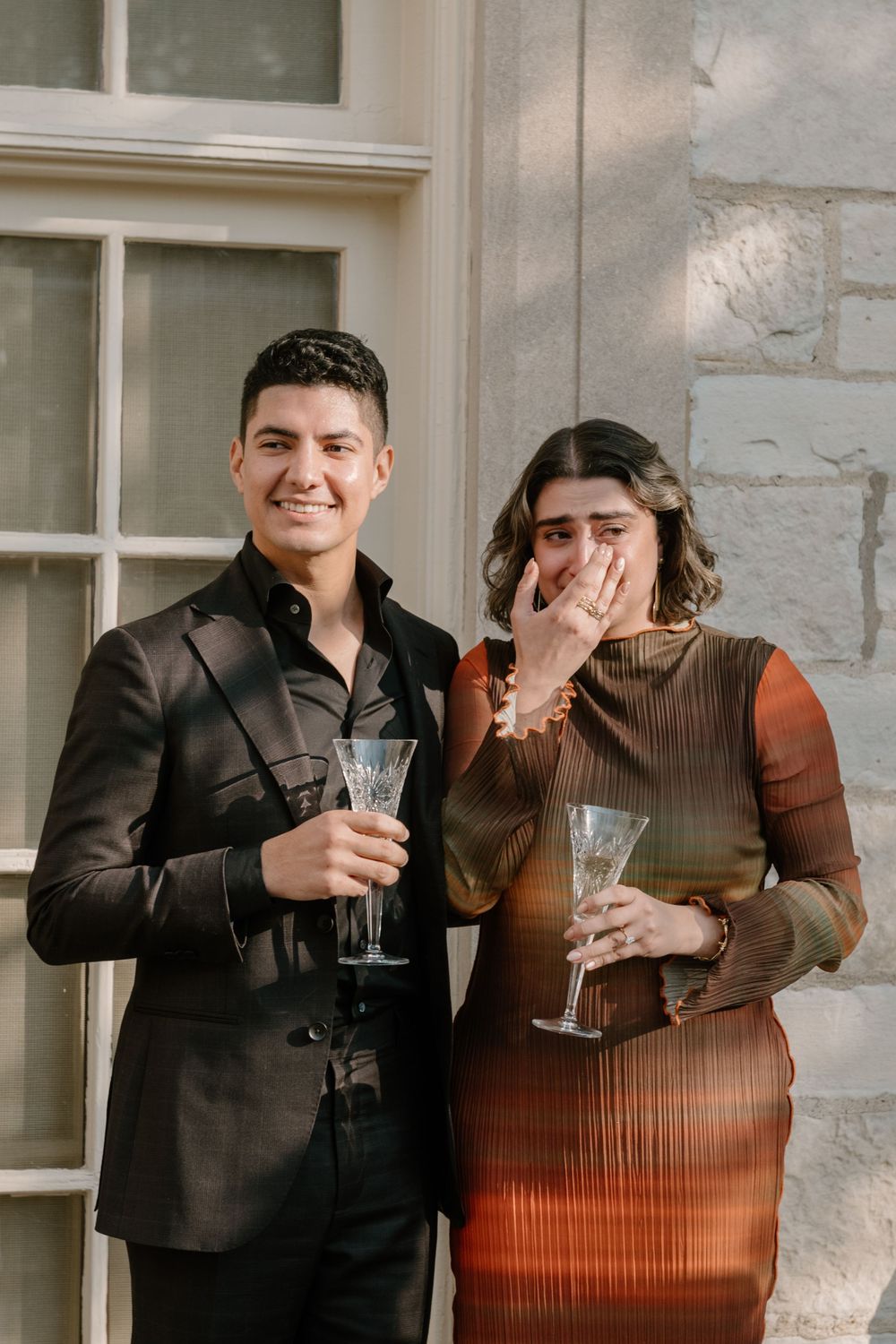 A couple in formal attire celebrates with champagne glasses while standing against a stone wall.