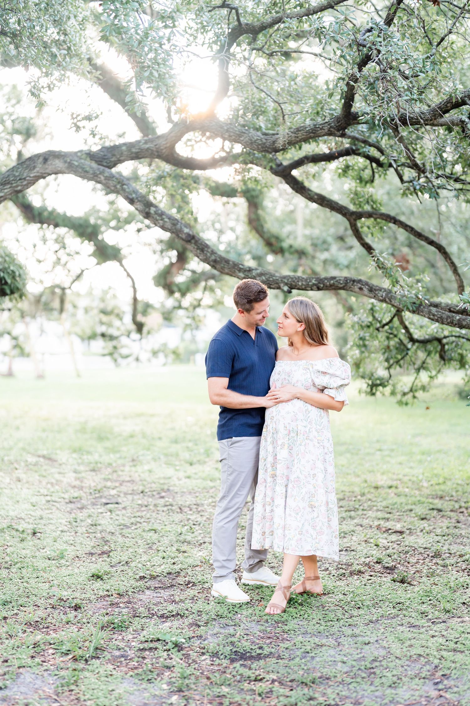 A couple stands together on a grassy hill with sunlit trees in the background during a maternity photo session.
